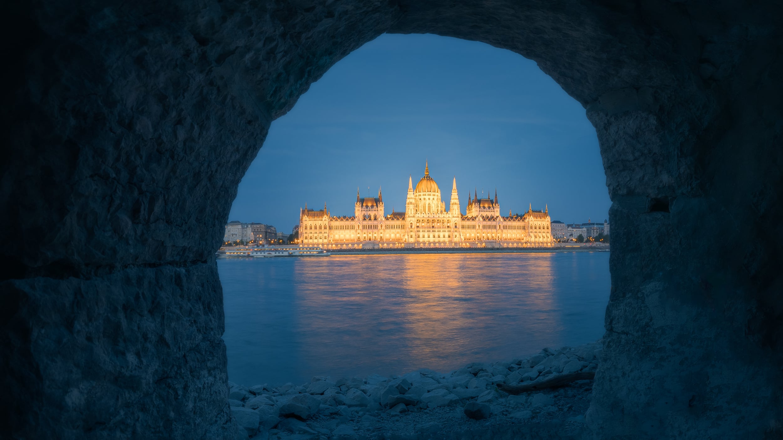 El Parlamento de Budapest iluminado en la hora azul, visto a través de un agujero en un muro de piedra junto al Danubio.